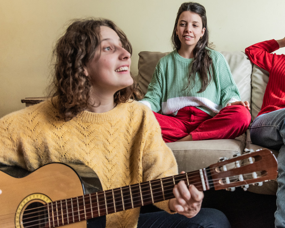 Zwei Personen sitzen auf einem Sofa, eine sitzt davor und spielt Gitarre.