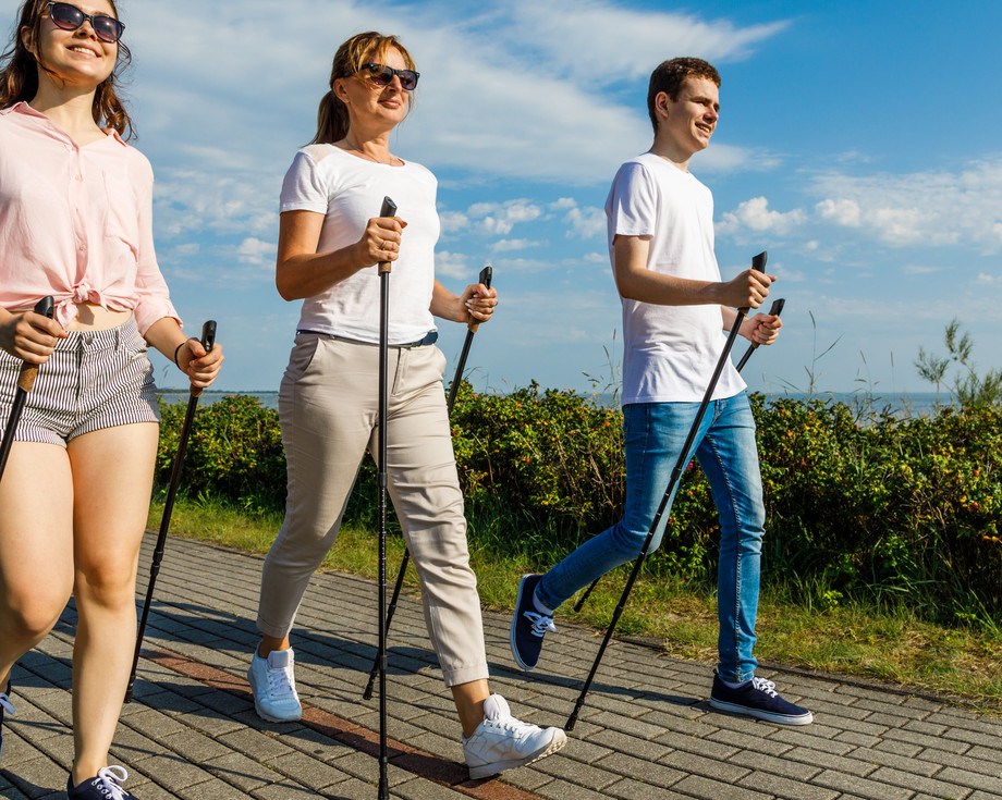 Drei Personen gehen mit Nordic Walking Stöcken auf einem Weg entlang, mit Blick auf den Ozean.