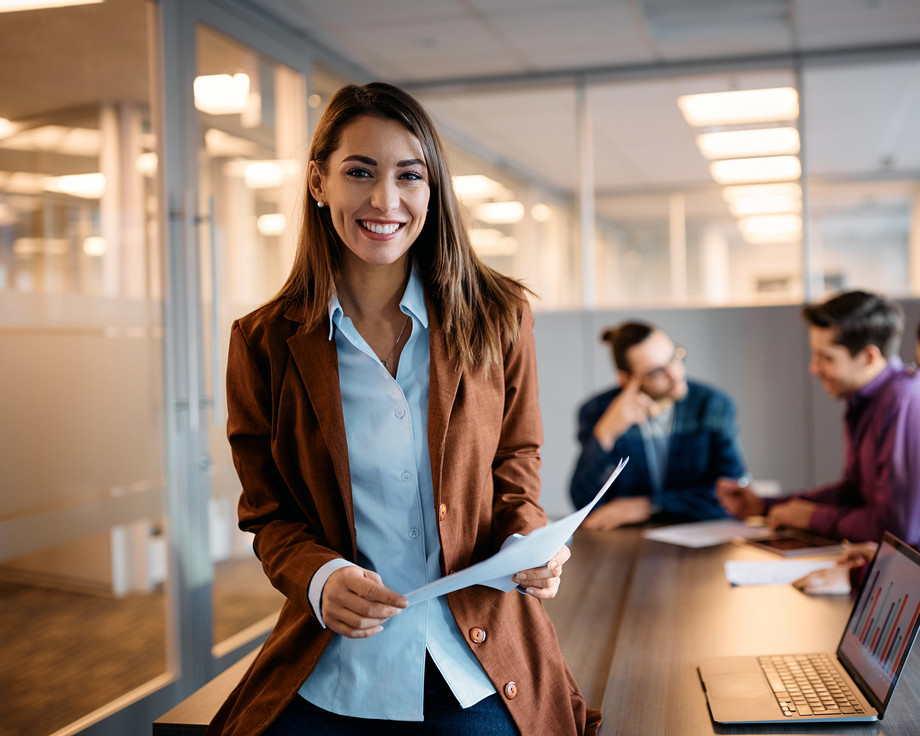 Eine Frau in einem Büro, die ein Dokument hält, während andere an einem Tisch arbeiten.