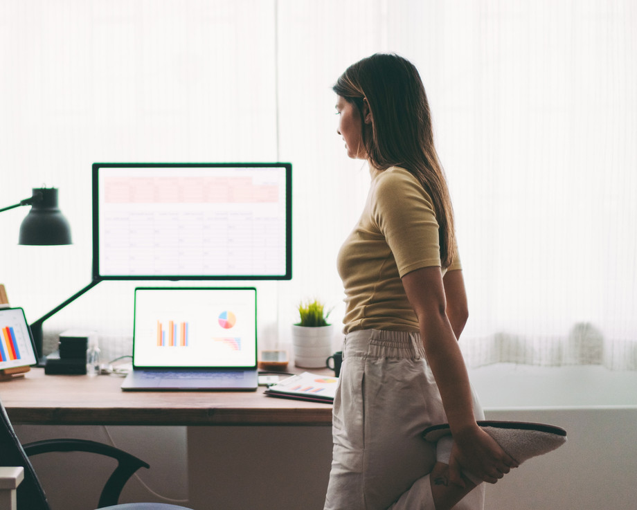 Frau steht an einem Schreibtisch mit Computer und Laptop.